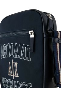 Borsa blu navy con una texture liscia, dotata di una tasca con zip, dettagli in argento e una tracolla in tessuto canvas con accenti a righe. Il telaio include il nome del marchio.
