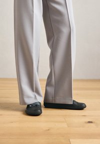 Ben med brede grå bukser og svarte slip-on sko med blå lisser står på et lyst tregulv.