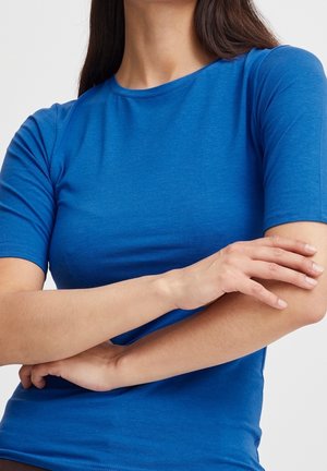 Kvinde iført en tætsiddende kortærmet blå skjorte med armene over kors foran en ensartet lys baggrund, med fokus på overkroppen og hænderne.