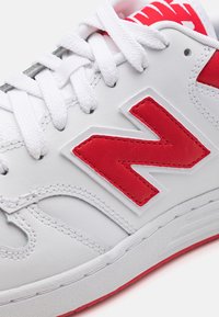 Vit syntetisk sneaker med röda accenter. Har en framträdande röd "N"-logotyp, texturerad yta och vita snören. Designen inkluderar sydda detaljer.