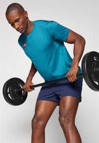 Teal sportshirt met een textuurafwerking en een oranje logo, gecombineerd met marineblauwe shorts. De persoon heft een zwarte halterstang.