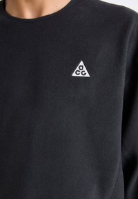Czarna bluza wykonana z miękkiego, teksturowanego materiału, z małym białym trójkątnym logo na piersi i okrągłym dekoltem.