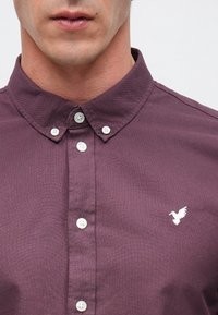 Homme portant une chemise prune boutonnée avec un petit logo oiseau blanc sur la poitrine et un col boutonné.