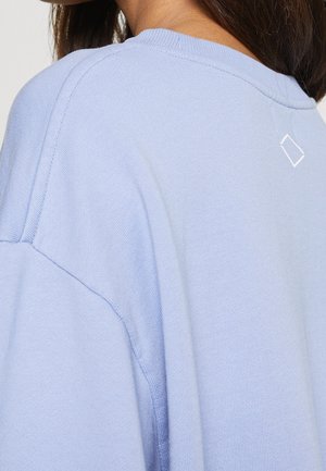 Zbliżenie na górną część pleców osoby noszącej jasnoniebieski sweter z małym białym logo w kształcie diamentu w pobliżu dekoltu.
