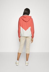 Chaqueta cortavientos en coral y blanco, con capucha, con un diseño en forma de cheurón en la espalda. Combinada con pantalones beige recortados y zapatillas deportivas.