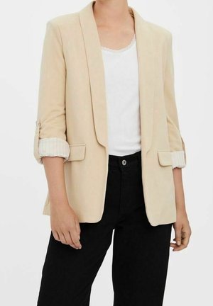 Femme portant un blazer beige avec les manches retroussées sur un haut blanc et un pantalon noir, debout devant un fond uni.
