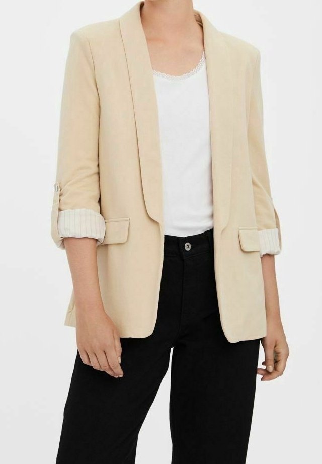 Femme portant un blazer beige avec les manches retroussées sur un haut blanc et un pantalon noir, debout devant un fond uni.