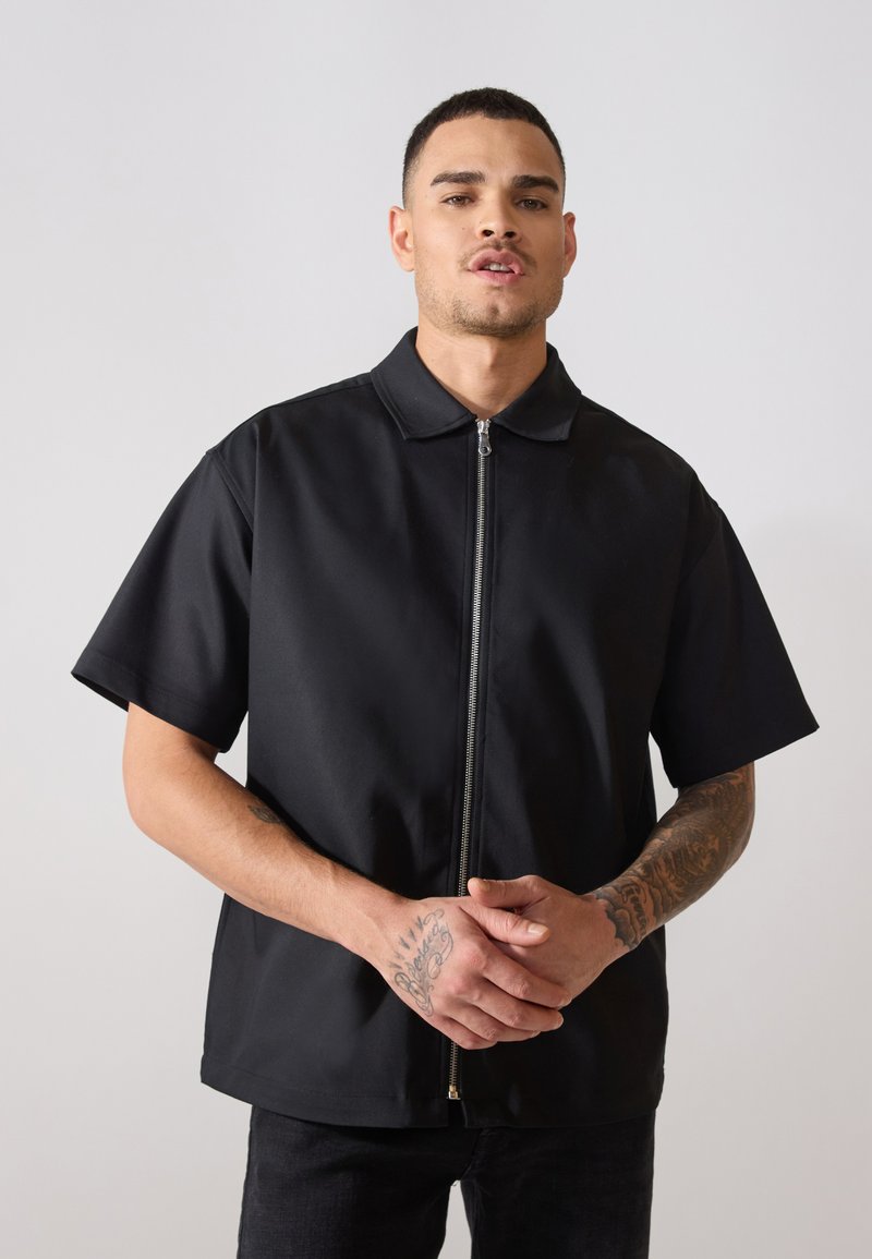 Μαύρο κοντομάνικο μπλουζάκι με φερμουάρ και γιακά, φτιαγμένο από λεία ύφασμα. Διαθέτει μπροστινό φερμουάρ και άνετη εφαρμογή. Απλός σχεδιασμός, χωρίς ορατά μοτίβα.