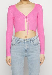 Rosa ribbad kort cardigan med långa ärmar, med V-ringning och knytning framtill. Bärs över ljusblå högmidjade jeans.