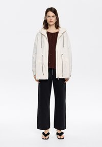 Helle beige Jacke mit Kordelzügen, Seitentaschen, über einem dunkelroten Strickpullover getragen. Schwarze Schlaghosen und schwarze Sandalen mit floralen Akzenten.