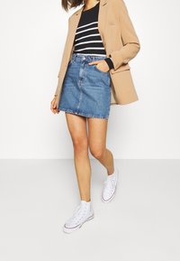Jeansrock in Hellblau, mit geradem Schnitt und genähten Details; kombiniert mit einem schwarz-weiß gestreiften Top und weißen Turnschuhen.