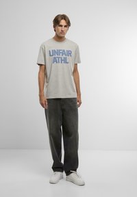 Šedé tričko s krátkým rukávem s modrým blokovým textem "UNFAIR ATHL" na přední straně, v kombinaci s tmavě šedými širokými kalhotami a bílými teniskami.