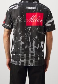 Czarna koszula z krótkim rękawem z wyrazistym czerwonym prostokątem z napisem "A.C. Milan", fakturowanymi wzorami graficznymi oraz kontrastującymi białymi detalami.
