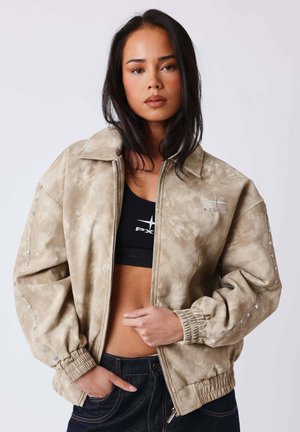 Jeune femme aux longs cheveux foncés portant une veste beige zippée par-dessus un crop top noir et un jean foncé, debout devant un fond uni.