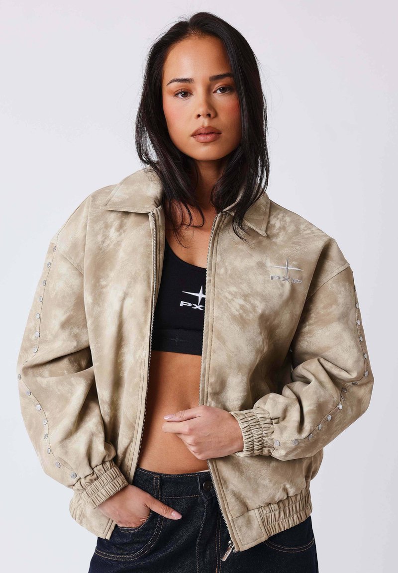 Jeune femme aux longs cheveux foncés portant une veste beige zippée par-dessus un crop top noir et un jean foncé, debout devant un fond uni.