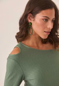 Top texturizado verde con escote en forma de barco y cortes en los hombros, combinado con grandes aros verdes que cuentan con acentos de cuentas.