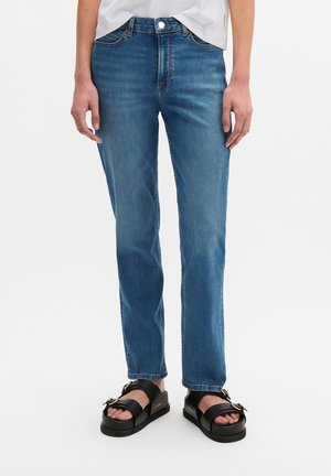 Persoon draagt blauwe jeans met rechte pijpen, wit topje en zwarte plateau sandalen met open tenen en gespen, staand op een effen witte achtergrond.