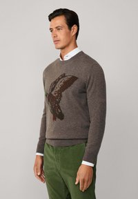 Maglione lavorato a maglia marrone con un design colorato di un'anatra che vola, colletto rotondo e orlo a costine, abbinato a pantaloni in corduroy verdi.