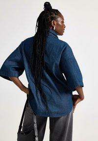 Denimskjorte i mørkeblå med løs pasform, brede ærmer og høj-lav kant. Har en spids krave og syet detaljer.