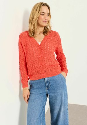 Žena v koralovom textúrovanom zapínacom kardigáne cez biely top a modré denimové džínsy, stojaca pri bielej stene s jednou rukou v vrecku.