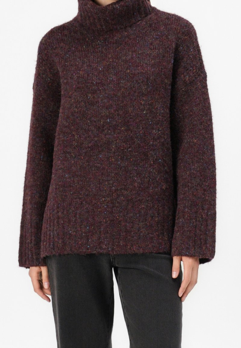 Femme portant un pull à col roulé texturé violet foncé avec des manches longues amples et un pantalon noir, debout devant un fond blanc uni.