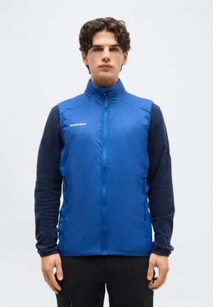 Νεαρός άνδρας που φοράει μπλε αμάνικο γιλέκο με φερμουάρ πάνω από μαύρο μακρυμάνικο πουκάμισο, στέκεται μπροστά σε απλό λευκό φόντο.