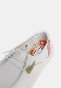 Witte canvas schoen met een gestructureerd oppervlak, crèmekleurige zool, decoratieve kwast en kleurrijk patroon aan de binnenkant. Heeft een eenvoudige vetersluiting.