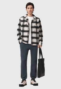 Czarna i biała koszula w kratę, biała koszulka, ciemne dresy, jasne sneakerzy, trzymająca czarną torbę na ramię. Swobodny, warstwowy strój.