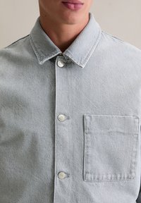 Hellgraues Denimhemd mit spitzem Kragen, silbernen Knopfverschlüssen und einer aufgesetzten Brusttasche. Verfügt über eine strukturierte Stoffoberfläche.