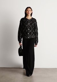Black sequined oversized sweater with a quilted pattern, paired with wide black trousers. Model holds a small black handbag. Smooth texture.