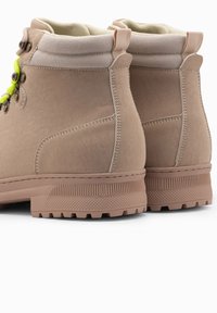 Botas de senderismo beige con parte superior de sintético texturizado, cuello acolchado en el tobillo, cordones en color verde lima y suela de goma resistente con un dibujo marcado.