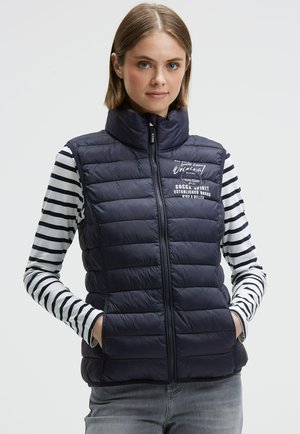 Navyblaue Steppweste mit hohem Kragen, Frontreißverschluss und gestepptem Textil. Getragen über einem schwarz-weiß gestreiften Langarmshirt.