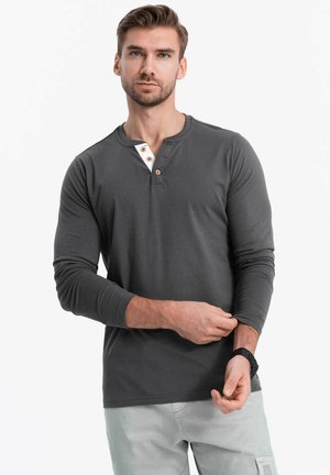 Szara, długosleeve koszulka henley z trzema guzikami na plastrze oraz białym akcentem przy dekolcie. Model w jasnych spodniach.