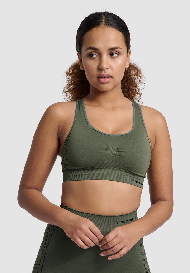 Vrouw met krullend haar in een groene sportbeha en bijpassende hoog getailleerde leggings, kijkt naar links, handen samengevouwen bij de taille.