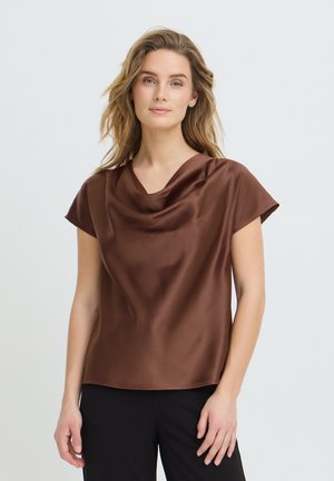 Naisel laineliste blondide juustega, pruunist satiinist lühikeste varrukatega pluusi ja mustade pükstega naine seisab tuhmi heleda tausta ees.