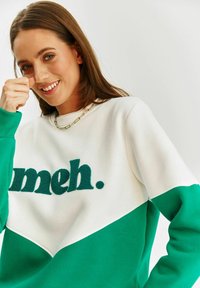 Bílý a zelený svetr s diagonálním designem; má texturu "meh." v tmavě zelené barvě přes hruď. Hladká látka s ribovanými manžetami.