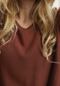 Haut couleur rouille en tissu texturé avec un décolleté arrondi. Manches longues et fluides ajoutent une coupe décontractée. Les cheveux encadrent partiellement le décolleté.