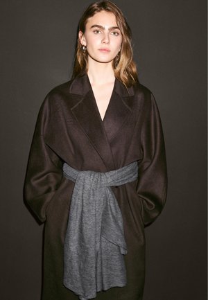 Cappotto oversized marrone scuro con maniche larghe, dotato di una cintura di stoffa grigia legata in vita. Texture liscia e colori a contrasto.