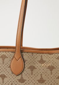 Ruskea olkalaukku, jossa on teksturoitu kangas ja logokuvio. Vankat vaaleanruskeat kahvat, joissa ommellut yksityiskohdat ja kultainen logokoriste.