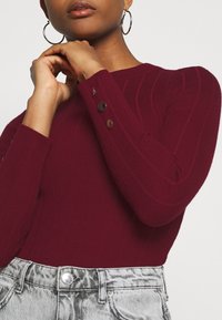 Pull en maille côtelée bordeaux à manches longues avec des boutons décoratifs aux poignets, assorti à un jean en denim argenté taille haute.