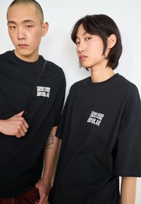 Two individuals wear oversized black t-shirts with a small white graphic on the chest. The fabric appears soft, with a relaxed fit.