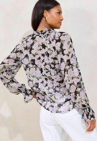 Transparente schwarze Bluse mit lila Blumenmustern, langen Ärmeln, gerafftem Ausschnitt und weit ausgestellten Manschetten. Kombiniert mit weißen Hosen.