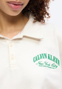 Κοντινό πλάνο ενός ατόμου που φοράει λευκό πόλο μπλουζάκι με πράσινο λογότυπο "Calvin Klein New York City" κεντημένο στο στήθος.