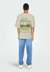 Beige übergroßes T-Shirt mit grüner Schrift und einem Palme-Grafik auf der Rückseite. Kombiniert mit hellblauen Jeans und weißen Sneakers.