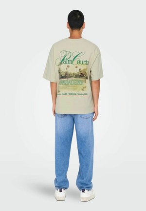 Beige übergroßes T-Shirt mit grüner Schrift und einem Palme-Grafik auf der Rückseite. Kombiniert mit hellblauen Jeans und weißen Sneakers.