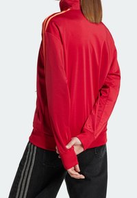 Röd träningsjacka med hög krage, långa ärmar och orangea ränder längs axlarna. Slät tyg med avslappnad passform.