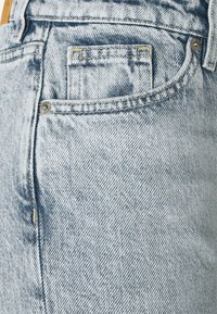 Ljusblå denimjeans med en texturerad yta. Har en liten framficka och gula sömnadsdetaljer, säkrade med metallnitar.