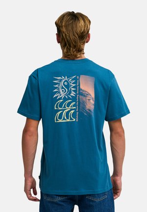 Uomo con acconciatura mullet che indossa una t-shirt blu con sole, onde e grafica fotografica sulla schiena, abbinata a jeans blu.