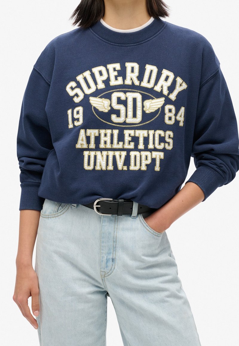 Personne portant un sweat-shirt bleu marine avec le texte "Superdry 1984 SD Athletics Univ Dpt", un jean bleu clair et une ceinture noire, une main dans la poche.