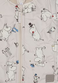 Svijetlosmeđi quiltani kaput s Moomin likovima u crnim konturama. Prednji zatvarač, rebrasti rubovi rukava i oznaka brenda pri dnu s desne strane.